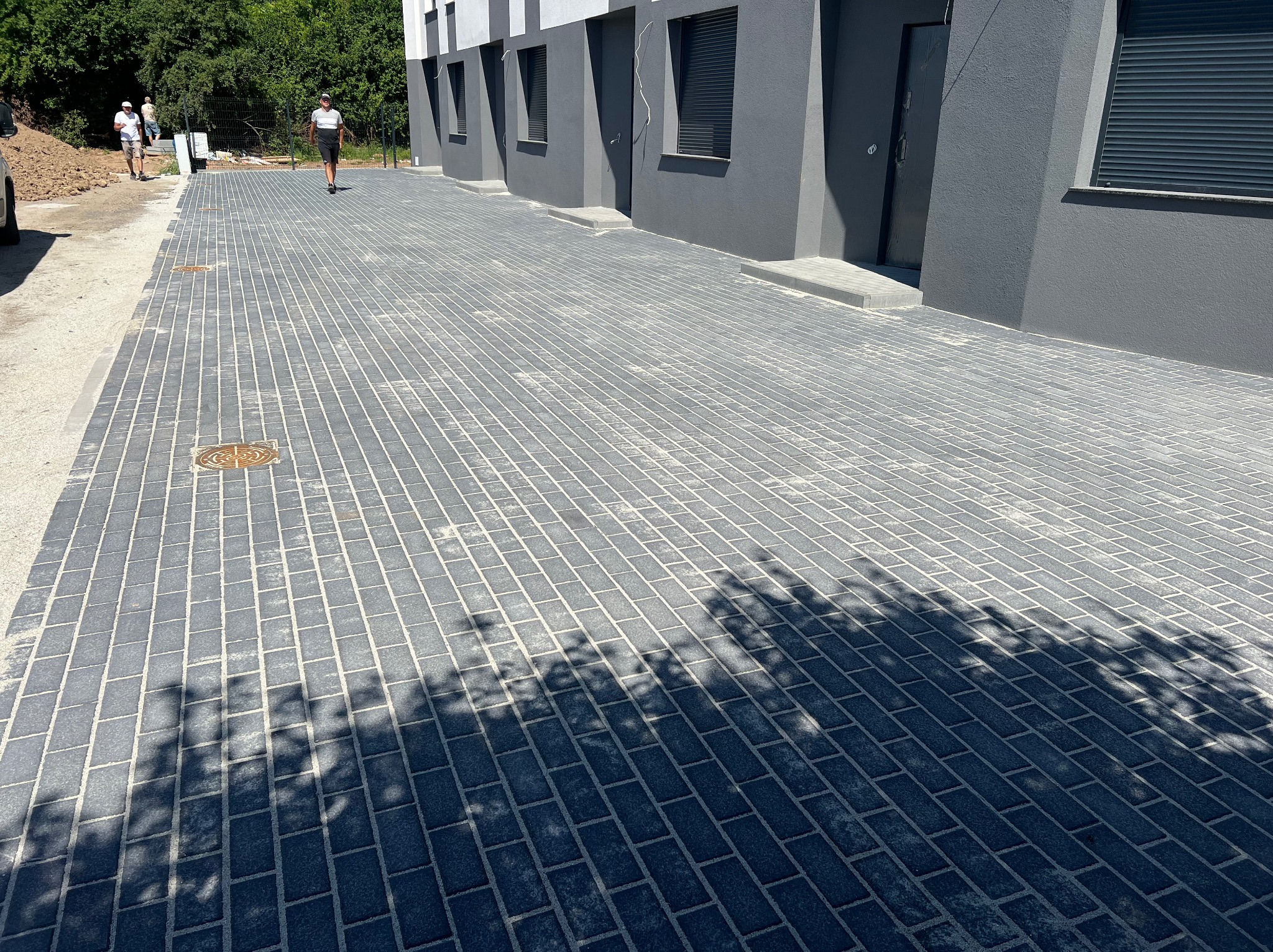 Ułożona, szara kostka brukowa przed budynkiem z ciemnymi oknami i drzwiami. Na kostce cień drzewa. W tle dwie osoby i częściowo widoczny samochód.