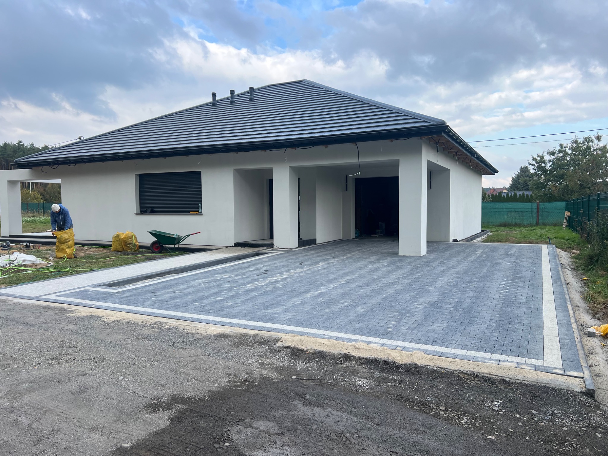Nowoczesny podjazd z szarej kostki brukowej, obrzeża z jasnej kostki, garaż dwustanowiskowy w tle, częściowo widoczny pracownik z taczką.