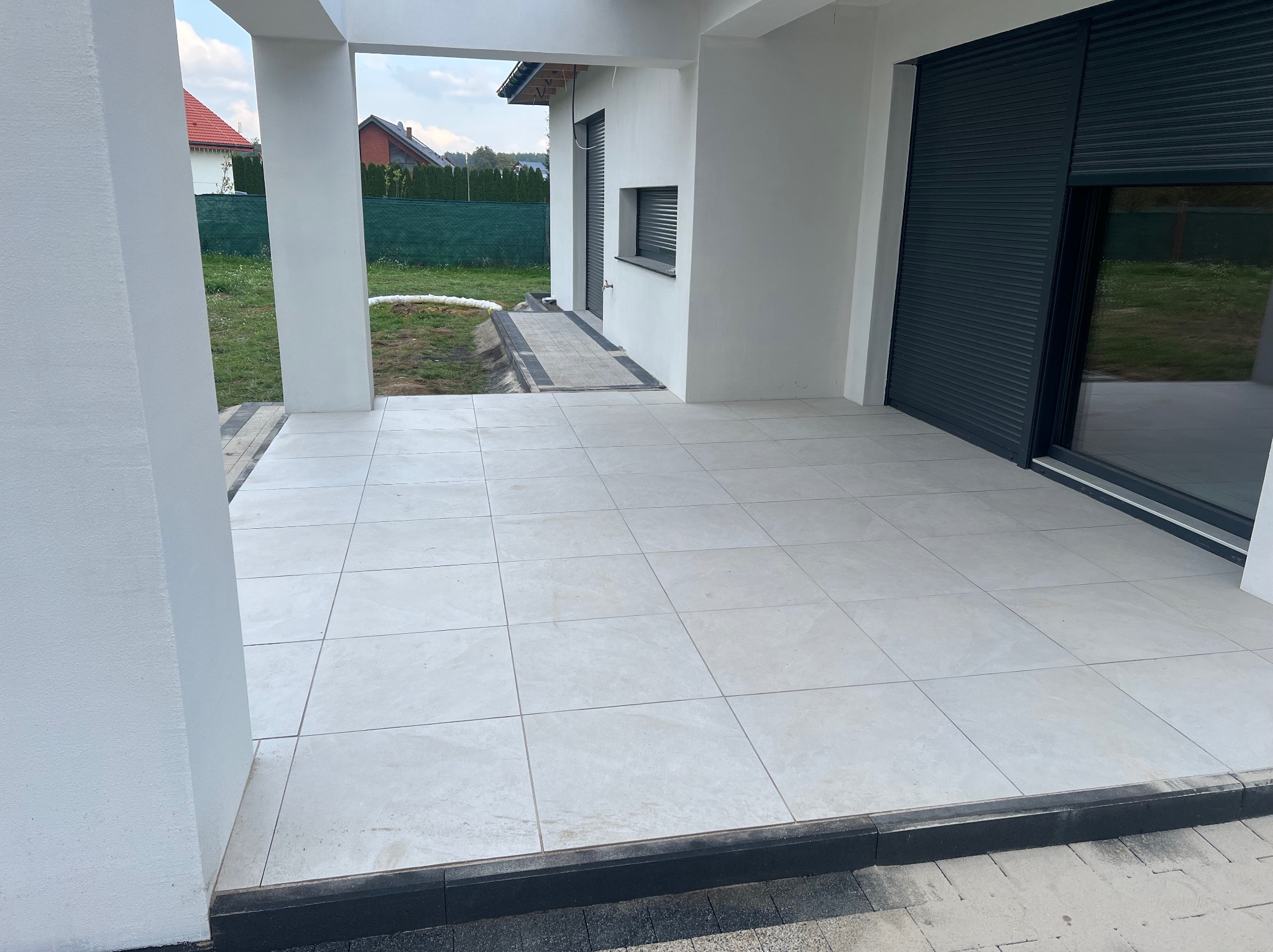 Wyłożony jasnymi płytkami taras przy nowoczesnym domu z szarymi roletami okiennymi. Widoczny fragment ogrodu i kostka brukowa na podjeździe.