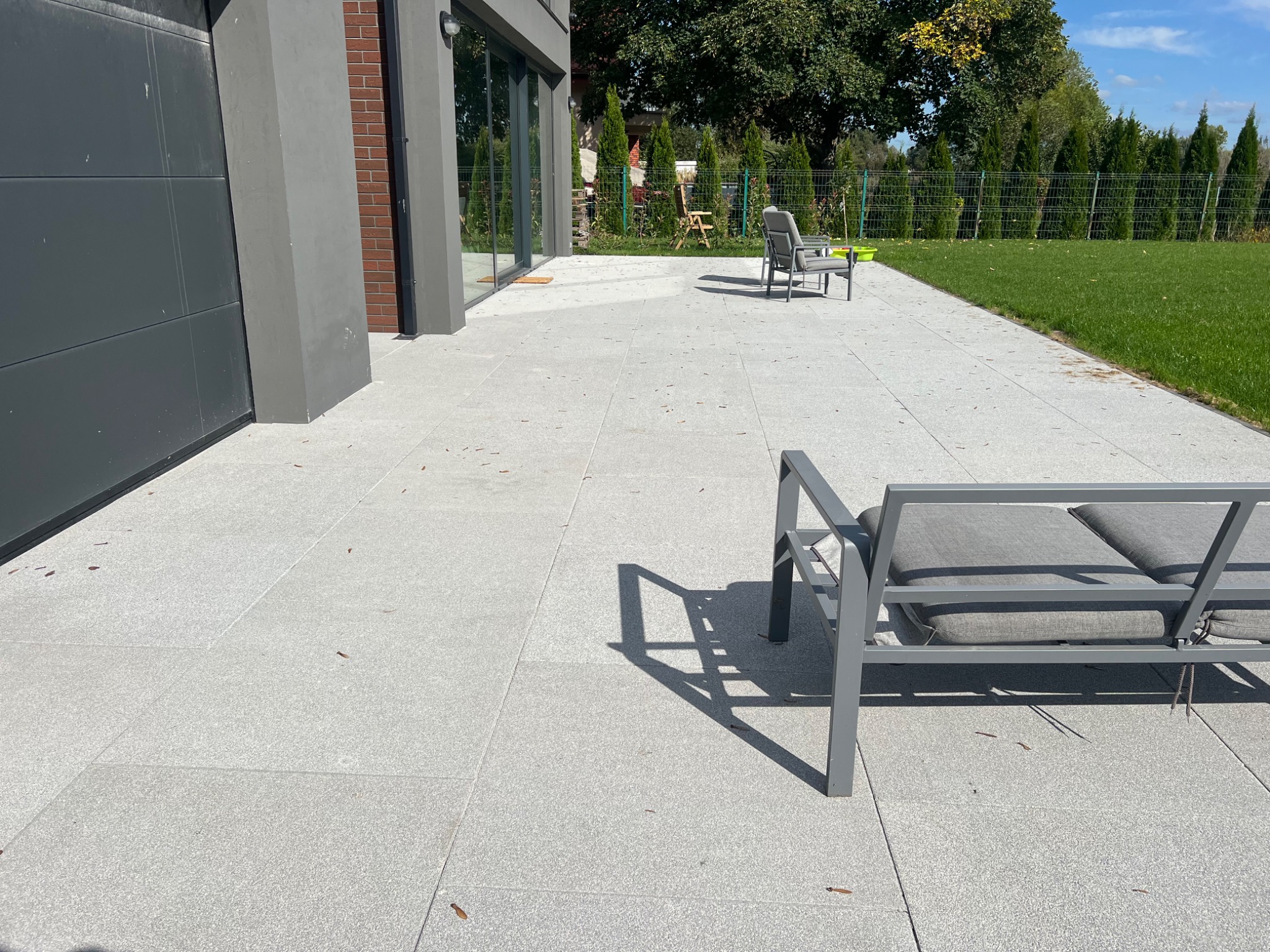 Jasnoszary taras z dużych płyt, z nowoczesnymi meblami ogrodowymi i widokiem na zielony trawnik za metalowym ogrodzeniem.