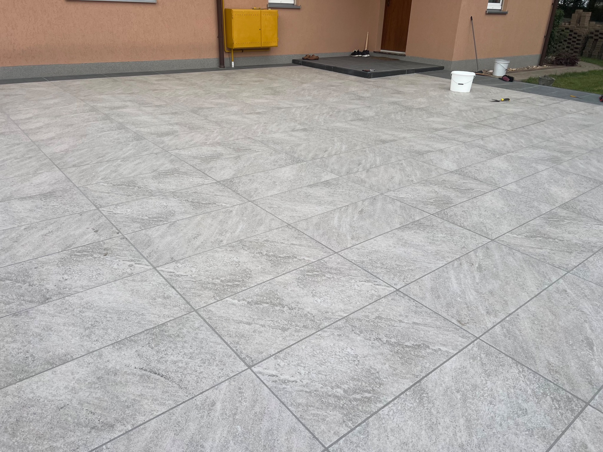 Jasnoszara, kwadratowa kostka brukowa ułożona pod kątem na podwórku domu. Widoczny fragment elewacji, drzwi wejściowe i narzędzia pracy.