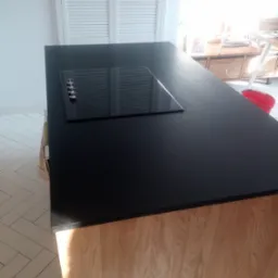 Czarny blat kamienny z płytą indukcyjną na wyspie kuchennej z drewnianym wykończeniem, widok z perspektywy, jasne wnętrze.