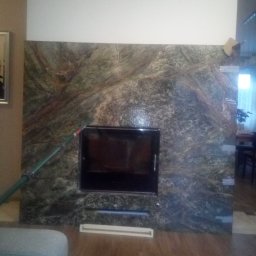 Albert Szymański - Nowoczesny kominek z obudową z zielonego marmuru, z czarnym wkładem i elementami wentylacyjnymi w salonie. W tle widoczny fragment wnętrza.