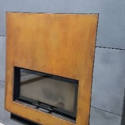 Albert Szymański - Nowoczesny kominek z metalową obudową w odcieniach rdzy i szarości, widoczny fragment szyby paleniska. Minimalistyczny design.