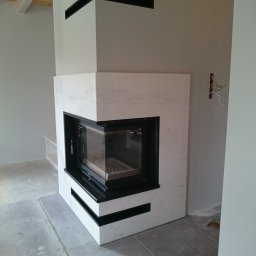 Albert Szymański - Nowoczesny kominek narożny z marmurową obudową i czarnymi akcentami, widoczny w surowym wnętrzu z odsłoniętymi przewodami elektrycznymi na ścianie.