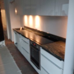 Albert Szymański - Nowoczesna kuchnia z białymi szafkami i blatem kamiennym w odcieniach brązu, oświetlona punktowymi lampami. Widoczny piekarnik.