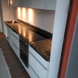 Albert Szymański - Kuchnia z blatem z ciemnego kamienia o wyraźnym użyleniu, wbudowaną płytą grzewczą i zlewozmywakiem. Białe szafki z poziomymi uchwytami. Oświetlenie podszafkowe.