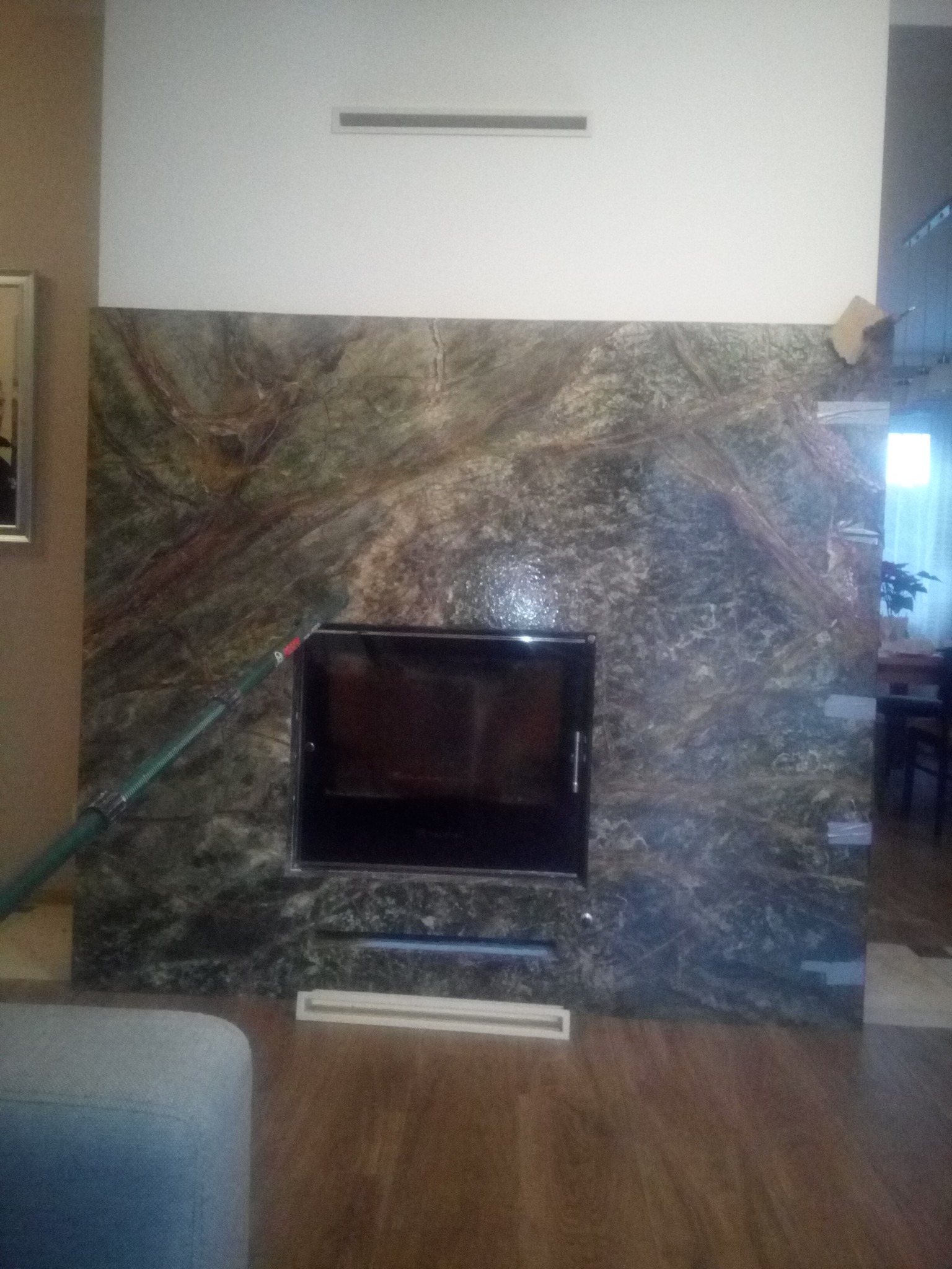 Nowoczesny kominek z obudową z zielonego marmuru, z czarnym wkładem i elementami wentylacyjnymi w salonie. W tle widoczny fragment wnętrza.