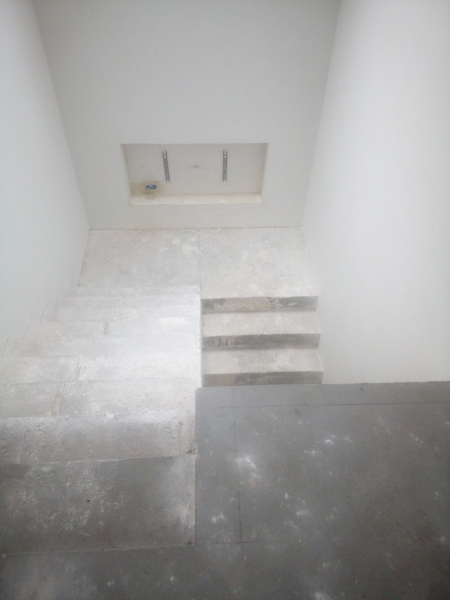 Widok z góry na schody w trakcie remontu, z szarymi stopniami i białymi ścianami. Widoczny otwór w suficie z elementami montażowymi.