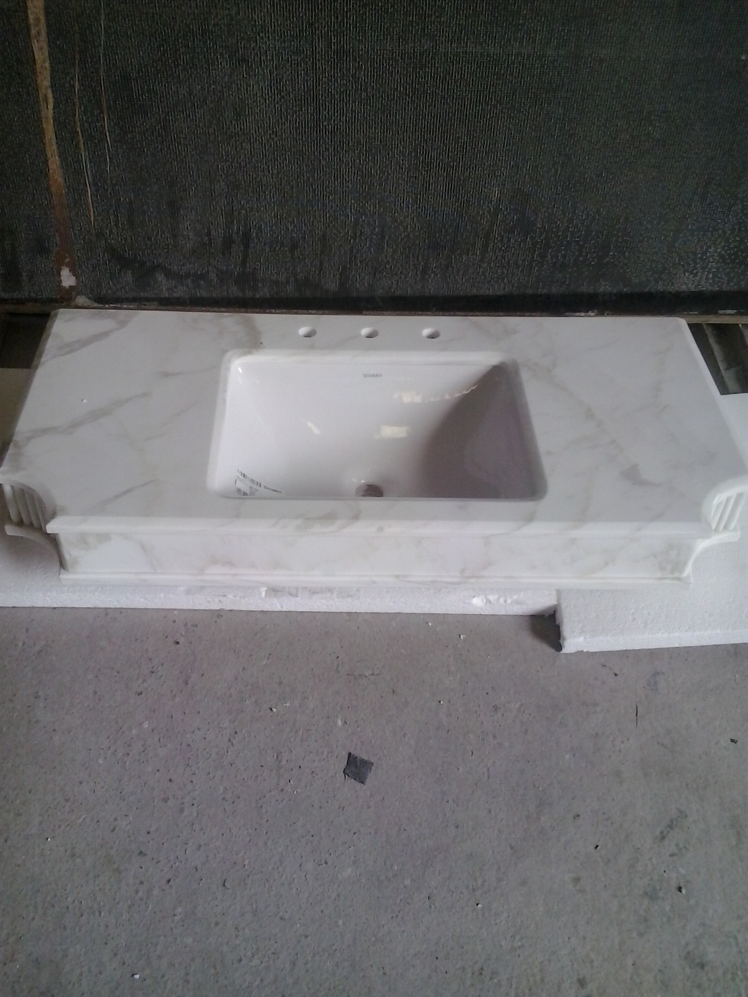 Biały blat kamienny z umywalką, imitujący marmur, z trzema otworami na armaturę. Umywalka ceramiczna, prostokątna. Tło stanowi szara ściana.