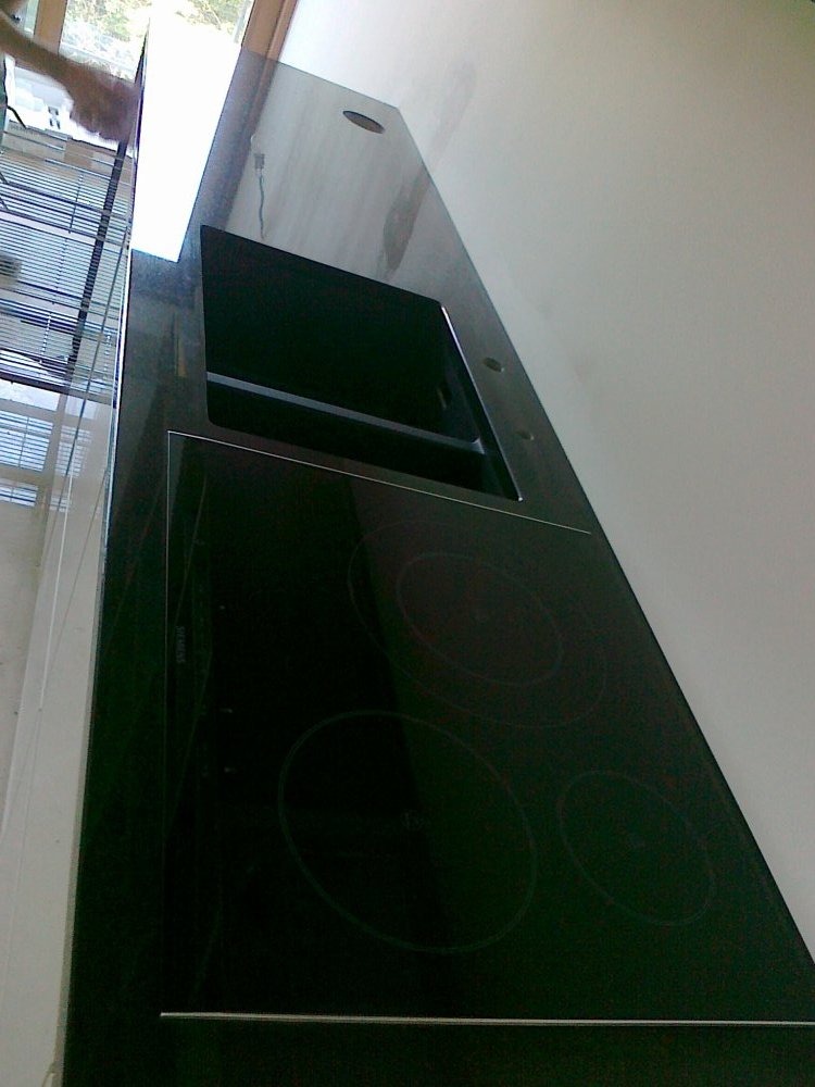 Czarny, błyszczący blat kamienny z wbudowaną płytą indukcyjną i zlewem, widoczny pod kątem, z fragmentem okna w tle. Minimalistyczny design, nowoczesna kuchnia.