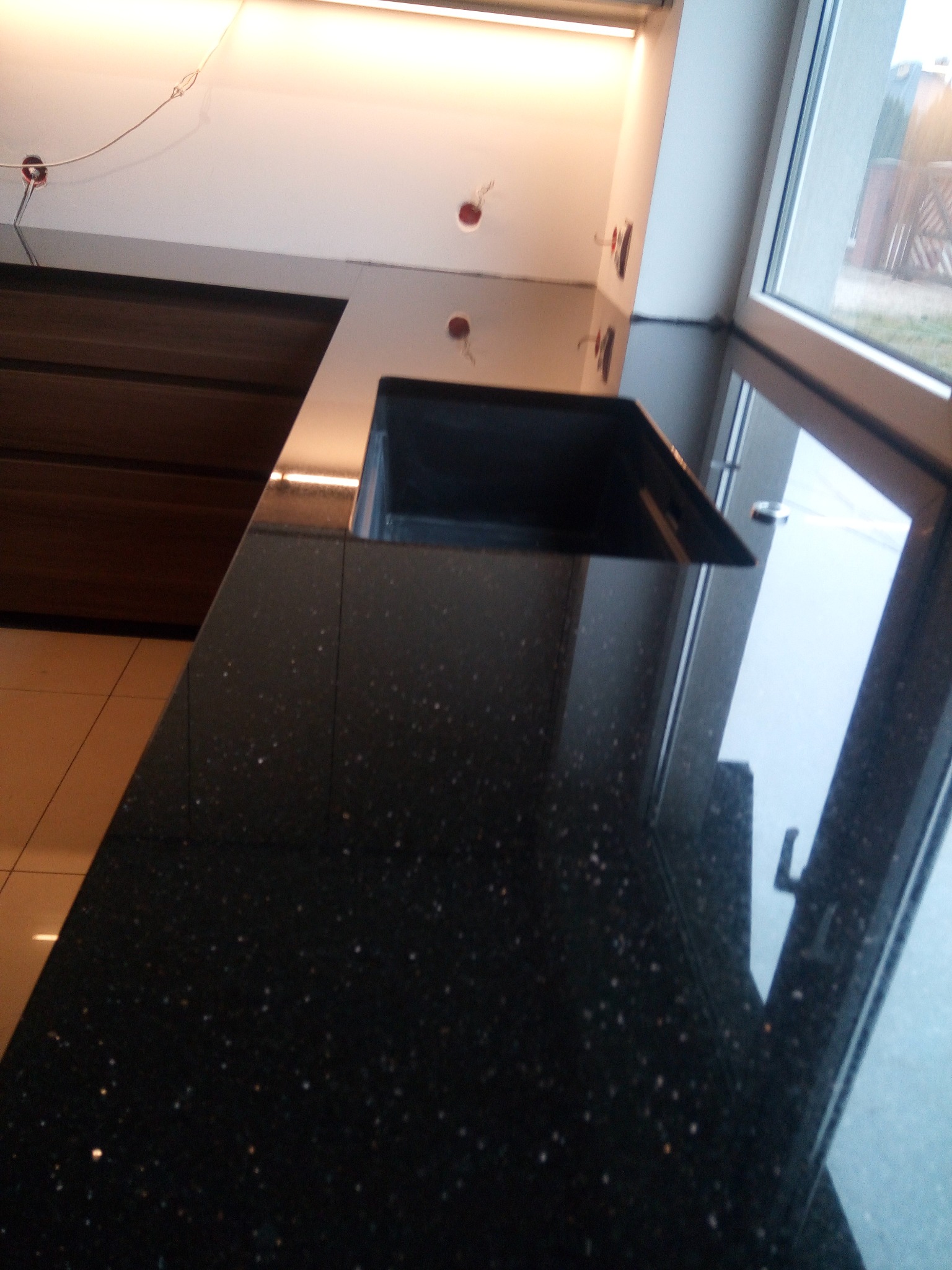 Błyszczący, czarny blat kamienny w kuchni, z wbudowanym zlewem, widoczny fragment okna i szafek, oświetlenie górne.