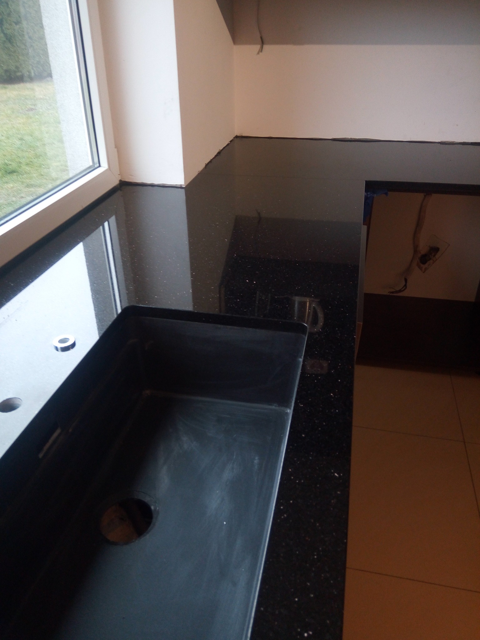 Błyszczący, czarny blat kamienny z brokatowym wzorem, obok okna i przygotowanym otworem na zlew, w trakcie montażu w kuchni.