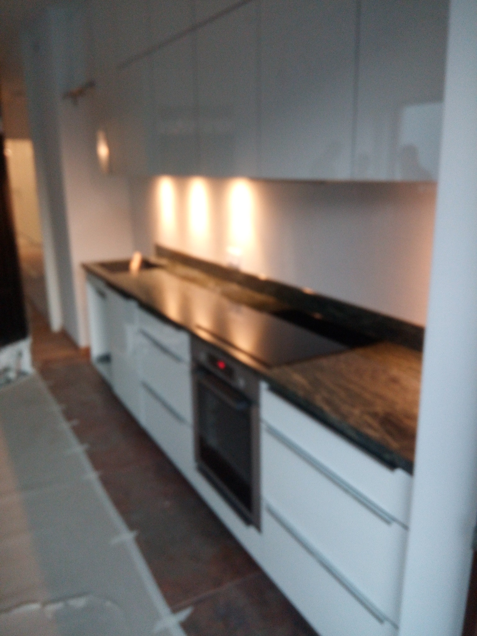 Nowoczesna kuchnia z białymi szafkami i blatem kamiennym w odcieniach brązu, oświetlona punktowymi lampami. Widoczny piekarnik.