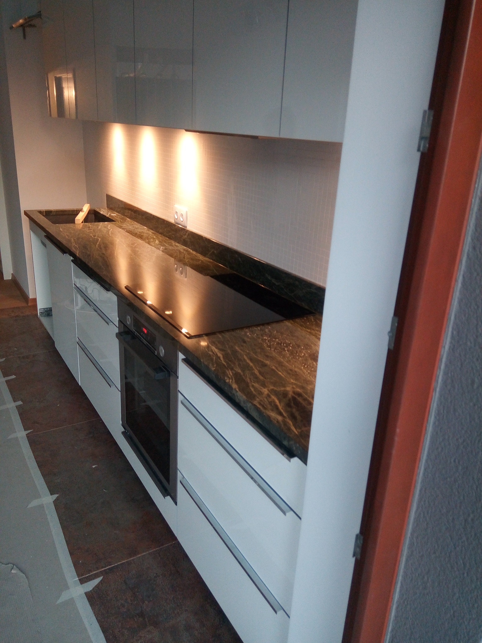 Kuchnia z blatem z ciemnego kamienia o wyraźnym użyleniu, wbudowaną płytą grzewczą i zlewozmywakiem. Białe szafki z poziomymi uchwytami. Oświetlenie podszafkowe.