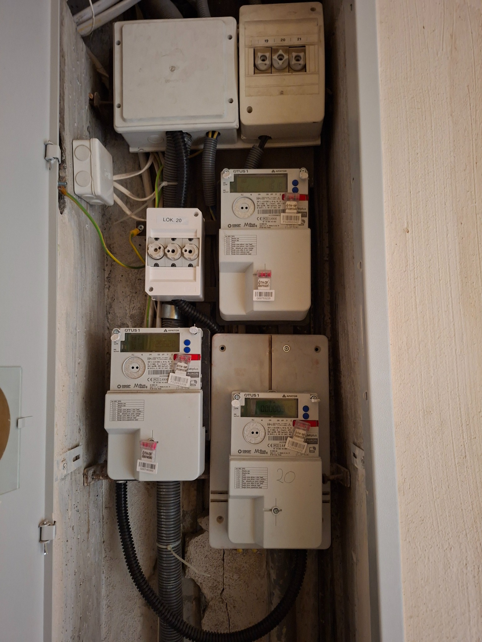 Skrzynka z czterema licznikami energii elektrycznej, zabezpieczeniami i przewodami w betonowej wnęce. Widoczne oznaczenia 'LOK. 20' i 'OTUS 1' na urządzeniach.