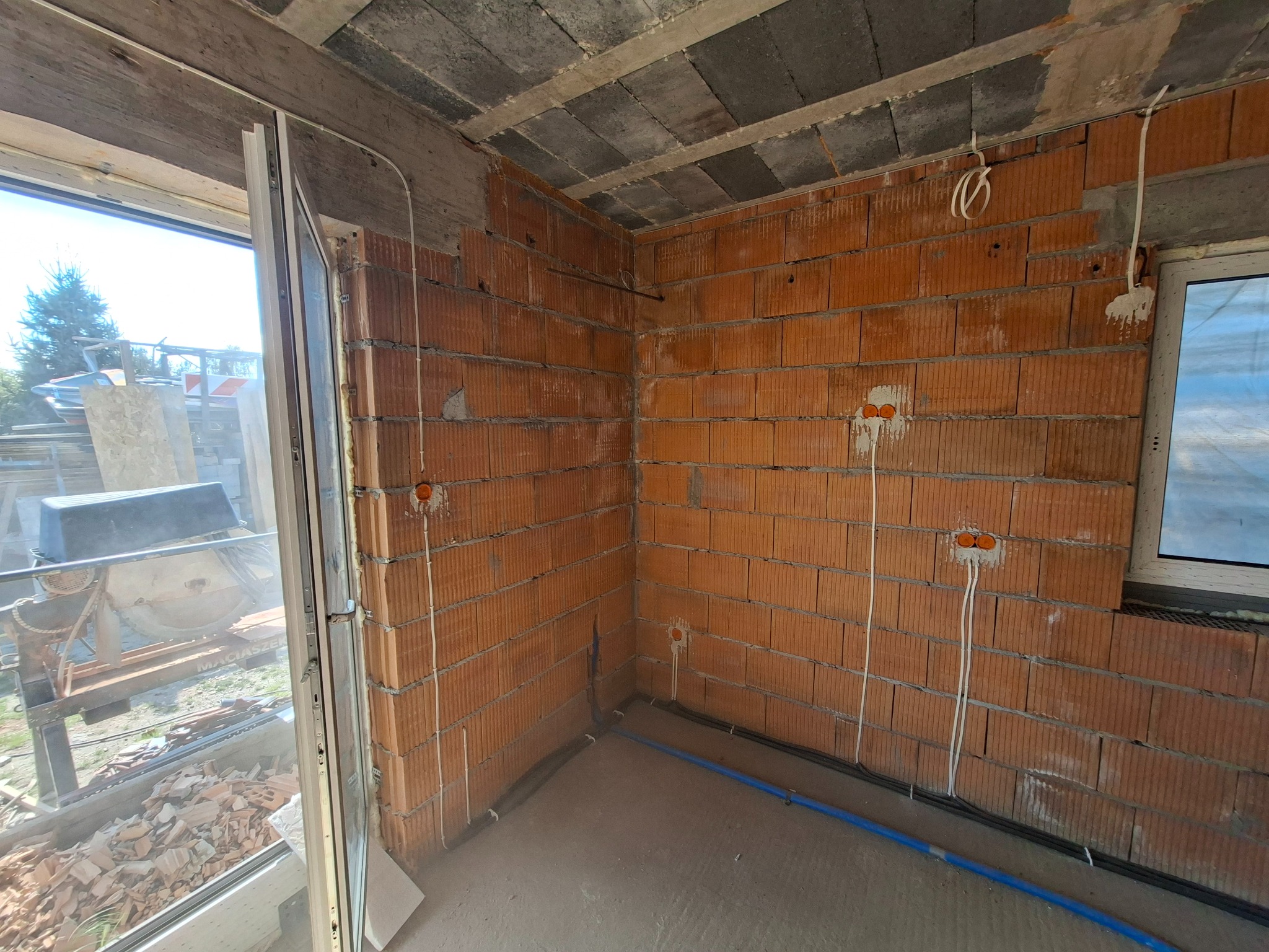 Surowy mur z instalacją elektryczną: kable w peszlu prowadzone pionowo wzdłuż ściany, widoczne puszki pod gniazdka. W tle maszyna do cięcia glazury.