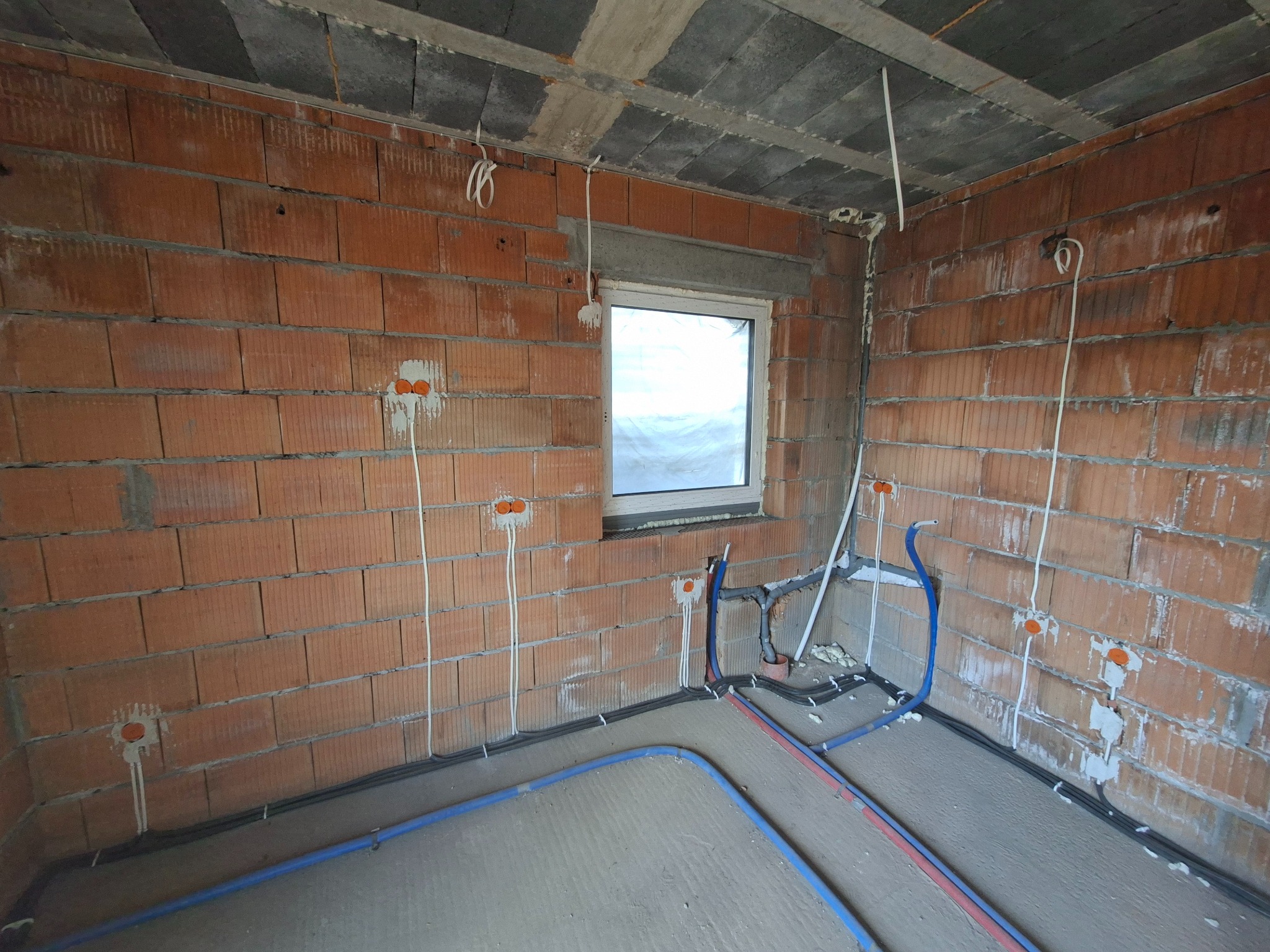 Surowy pokój z ceglanymi ścianami w trakcie instalacji elektrycznej i hydraulicznej. Widoczne przewody i rury w podłodze oraz puszki elektryczne w ścianach.