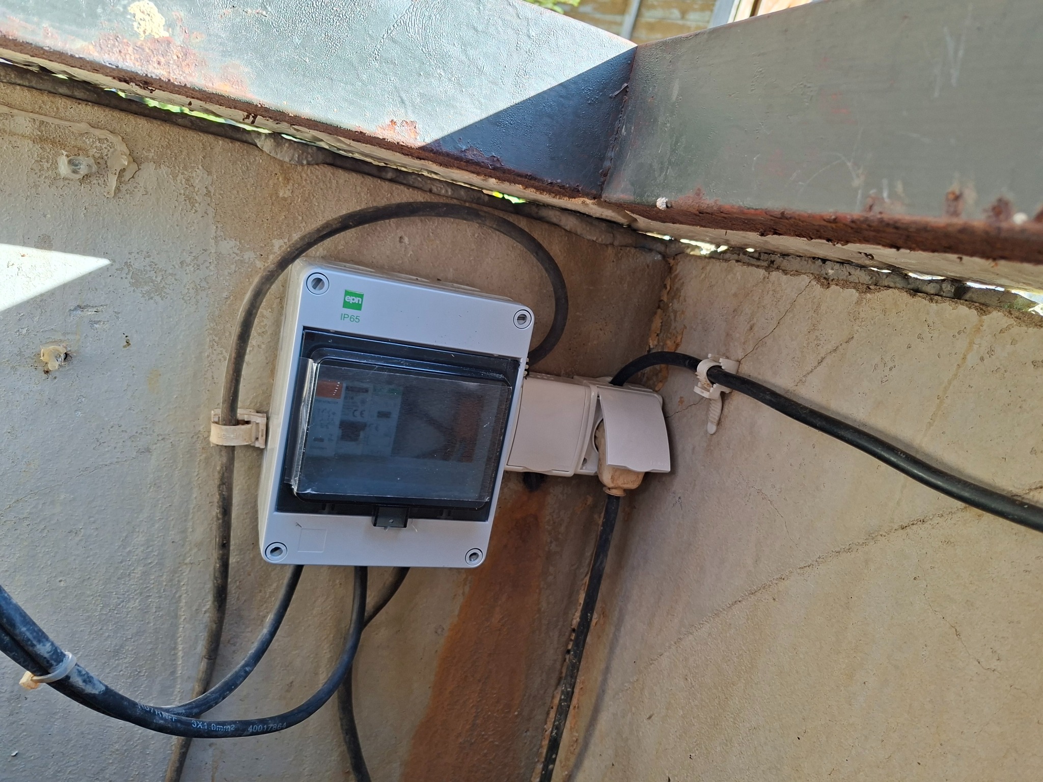 Szara skrzynka elektryczna IP65 z widocznymi bezpiecznikami, zamontowana w rogu betonowej konstrukcji, z podłączonymi kablami. Widoczne uchwyty mocujące przewody.