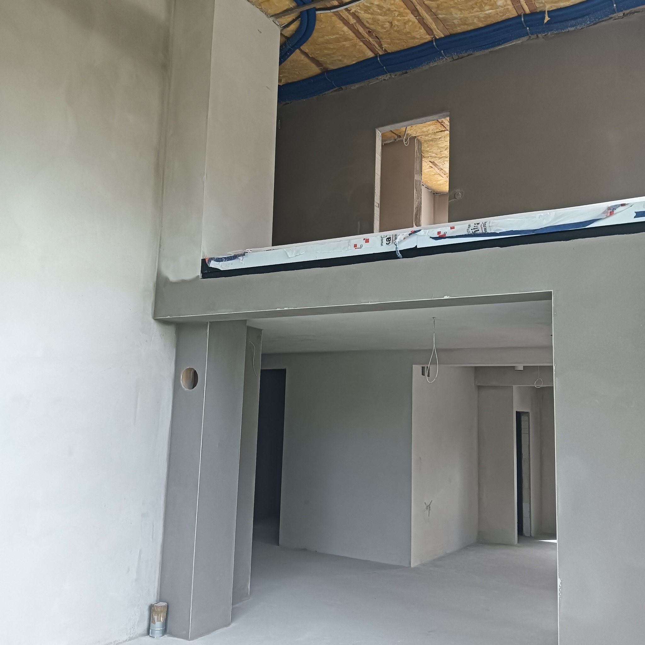 Surowe wnętrze po nałożeniu tynku, widoczne przygotowania do dalszych prac wykończeniowych. Instalacje pod sufitem, dwupoziomowa przestrzeń, szpachlowane ściany.