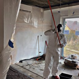 REMO GOR DOMINIK GÓRA - Pracownik w kombinezonie i masce maluje sufit wałkiem na długim kiju w pomieszczeniu zabezpieczonym folią malarską, widoczne czerwone buty i czarne wiaderko.