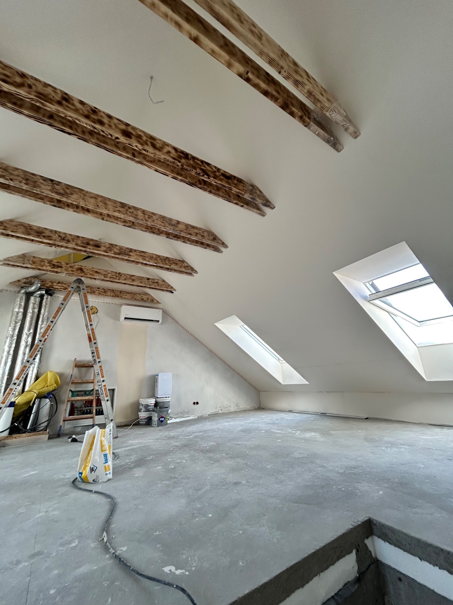 Poddasze w trakcie prac wykończeniowych: widoczne drewniane belki stropowe, okna dachowe, drabina, worki z zaprawą oraz ślady po szpachlowaniu na ścianach.