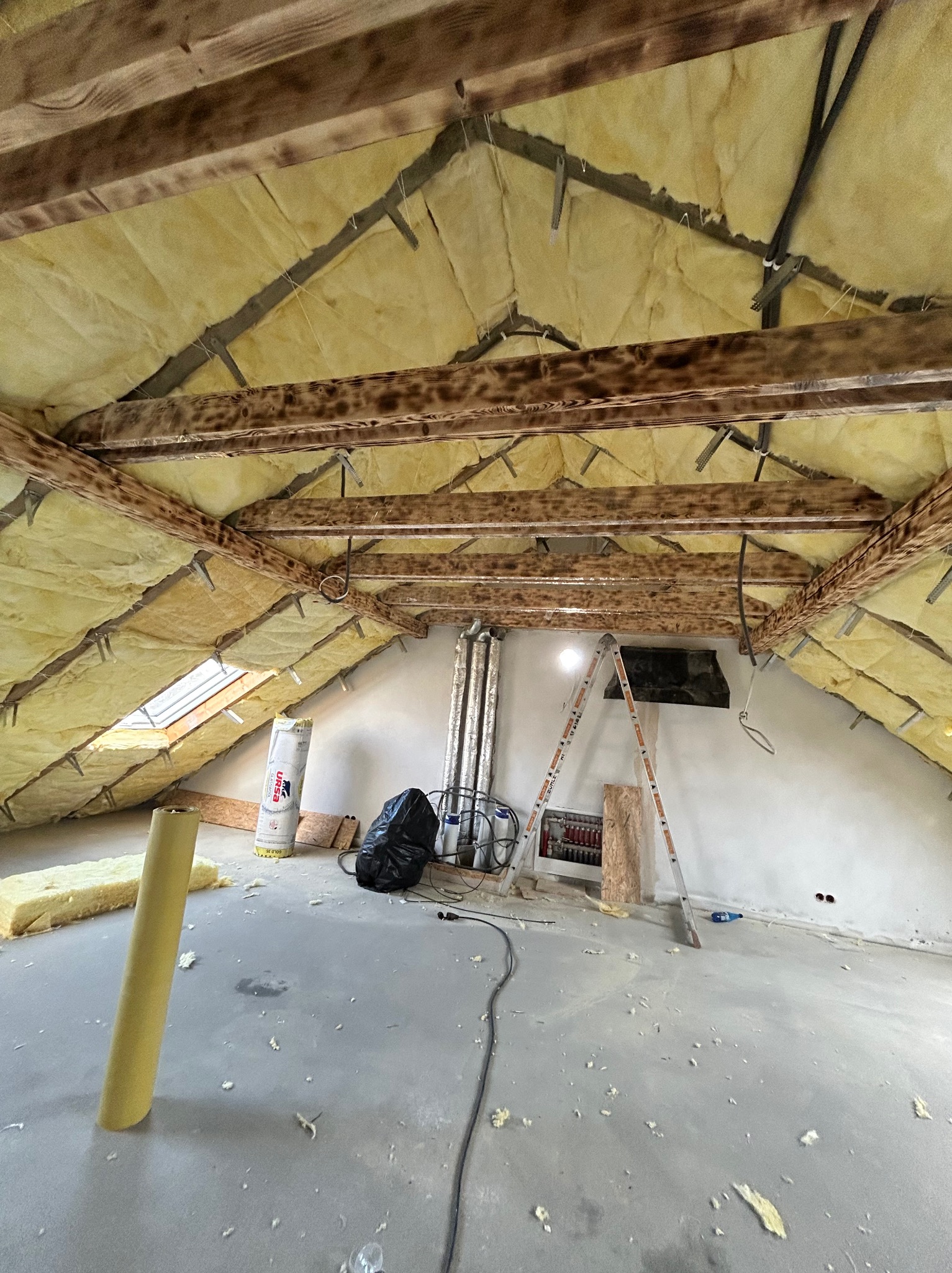 Poddasze w trakcie ocieplania wełną mineralną, widoczne drewniane belki stropowe, okno dachowe, drabina i materiały izolacyjne na podłodze.