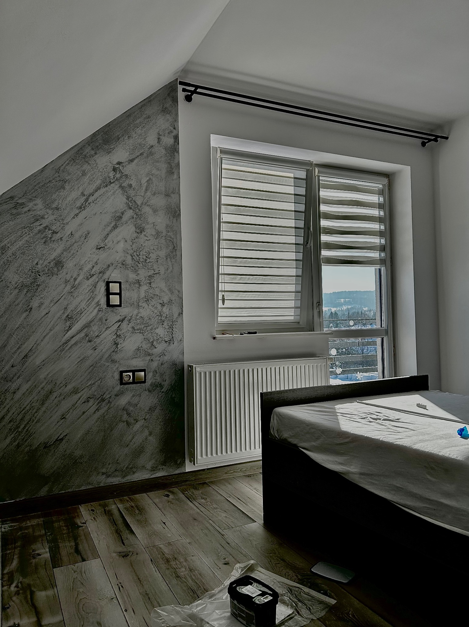 Szara ściana w sypialni z efektem dekoracyjnym przypominającym marmur, czarne włączniki światła, okno z żaluzjami dzień-noc, grzejnik i częściowo widoczne łóżko, na podłodze panele i akcesoria...