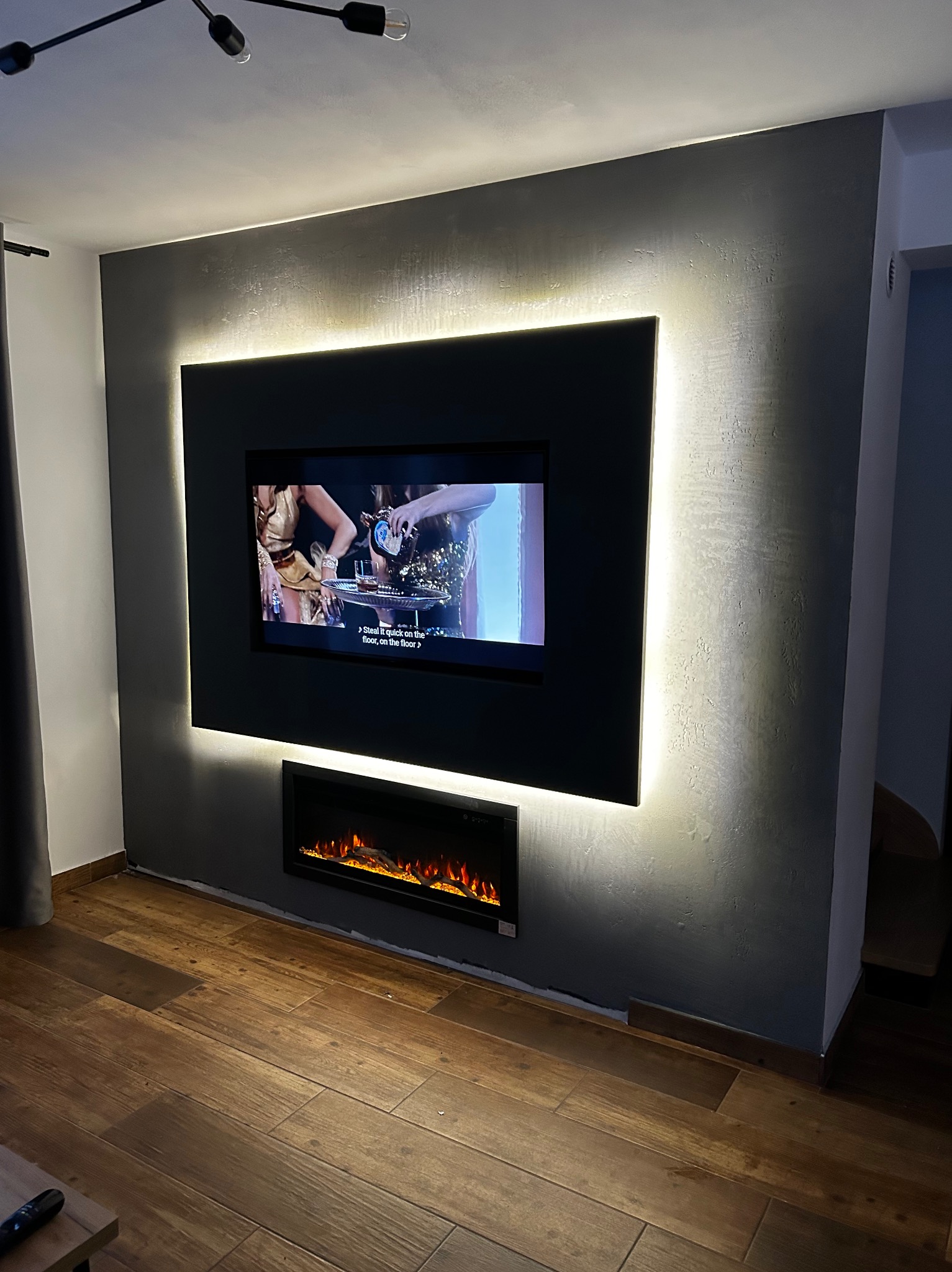 Telewizor w czarnej ramie z podświetleniem LED na szarej ścianie z dekoracyjnym tynkiem, pod nim elektryczny kominek, drewnopodobna podłoga.