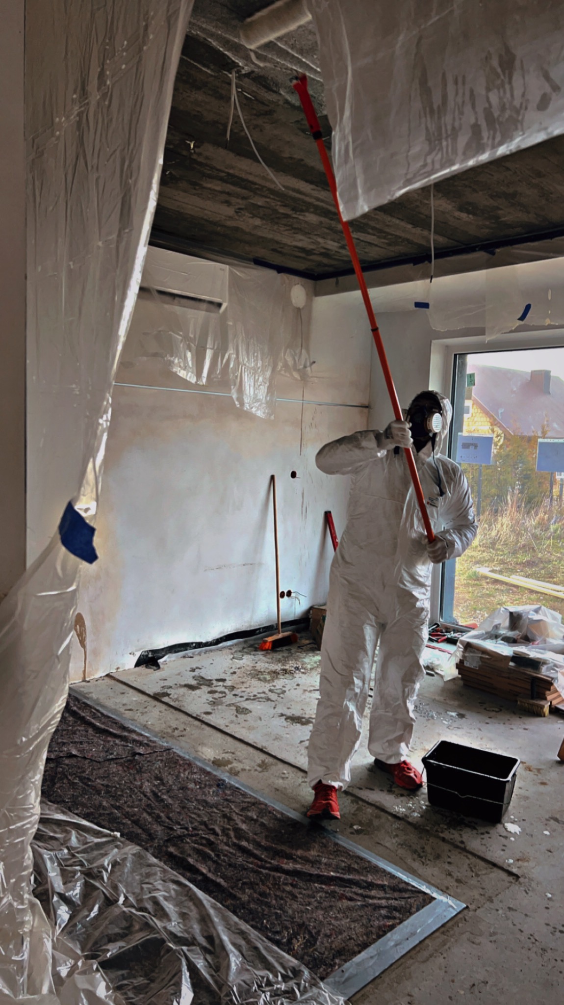 Pracownik w kombinezonie i masce maluje sufit wałkiem na długim kiju w pomieszczeniu zabezpieczonym folią malarską, widoczne czerwone buty i czarne wiaderko.