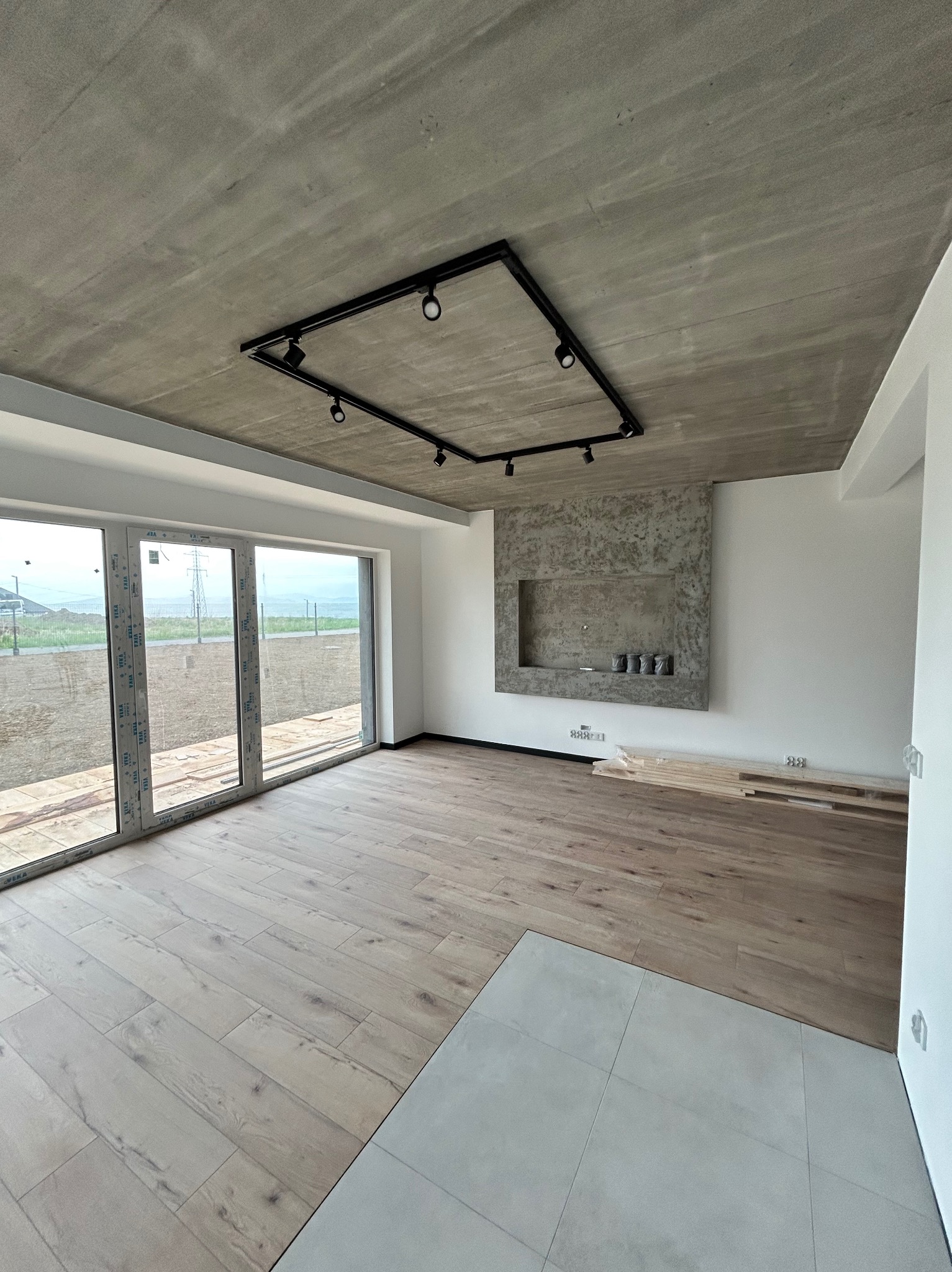 Jasne panele podłogowe w surowym wnętrzu z betonowym sufitem i ścianą z dekoracyjną wnęką. Widoczne duże okno i elementy instalacji elektrycznej.