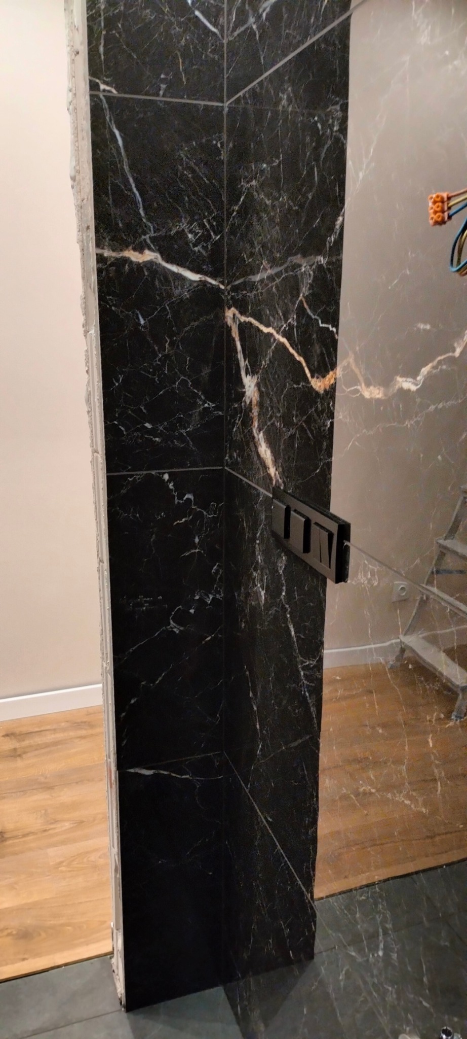 Częściowo wykończona kabina prysznicowa z czarnych płytek imitujących marmur, widoczne niedokończone fugowanie i instalacja szklanej ścianki, w tle drabina i podłączenie elektryczne.