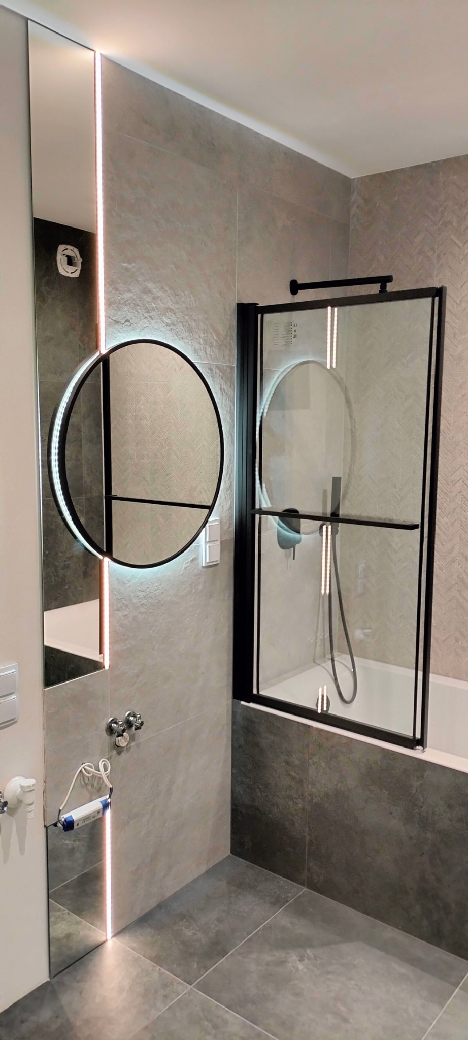 Nowoczesna łazienka z szarymi płytkami, okrągłym lustrem z podświetleniem LED i czarną kabiną prysznicową typu walk-in.
