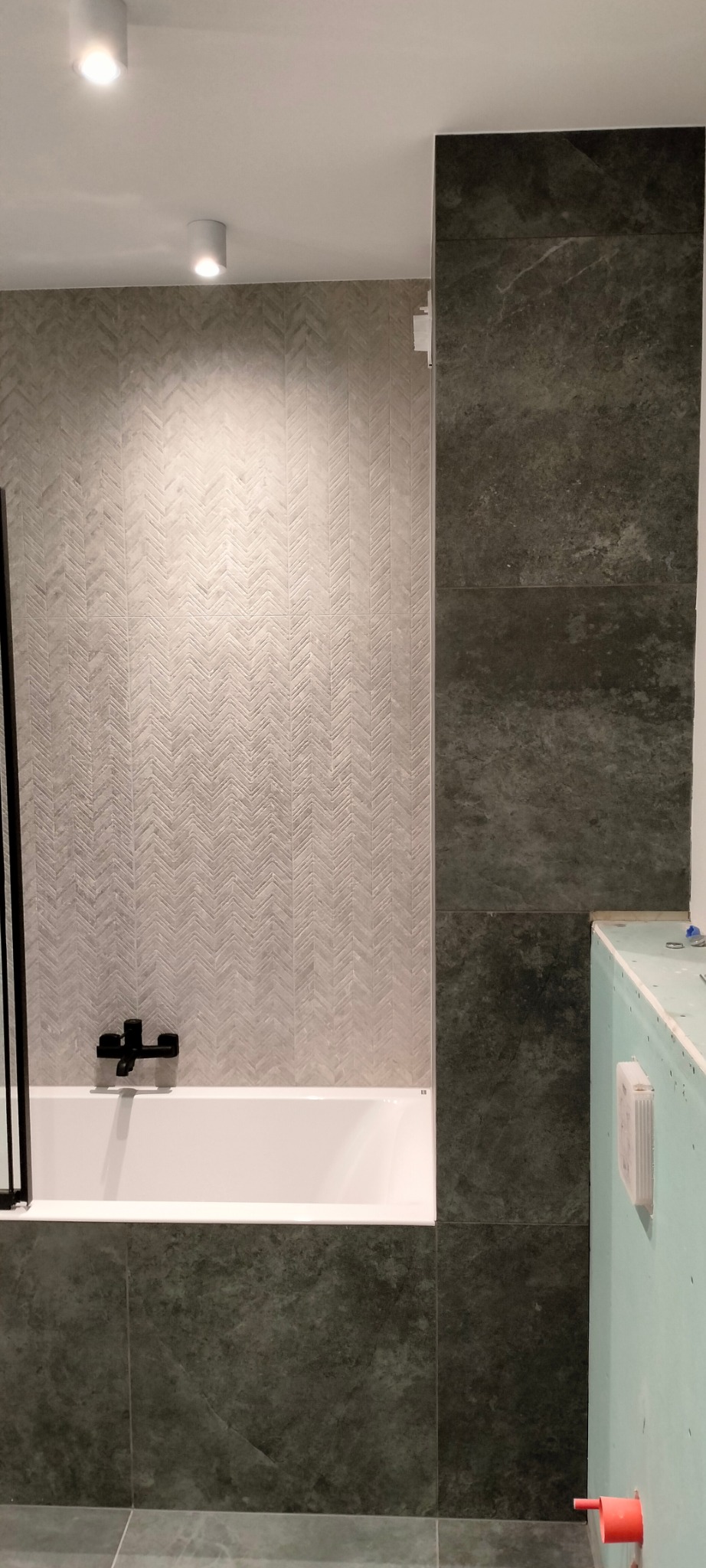 Fragment łazienki z wanną, baterią w kolorze czarnym i ścianą wyłożoną jasnymi płytkami w jodełkę oraz ciemnymi płytkami imitującymi kamień, widoczna instalacja wodna w ścianie obudowanej płytą...