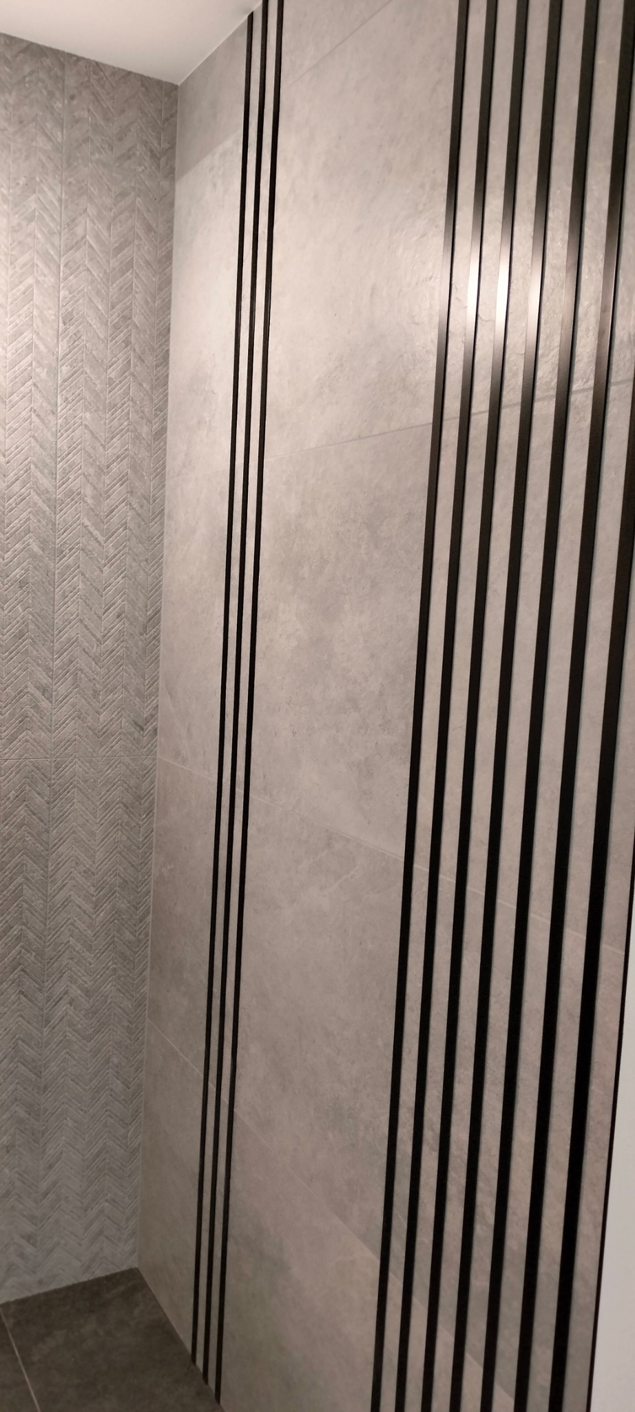 Ściana wykończona płytkami w odcieniach szarości, z dekoracyjnymi czarnymi listwami pionowymi; wzór jodełki na jednej ze ścian.