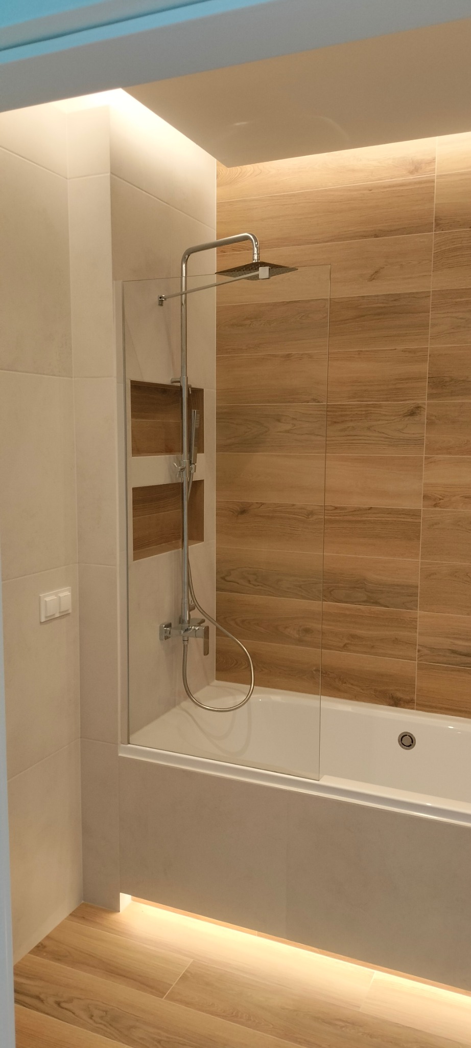 Nowoczesna łazienka z wanną, szklanym parawanem nawannowym, deszczownicą i drewnopodobnymi płytkami na ścianie oraz podświetleniem LED pod wanną i nad sufitem.