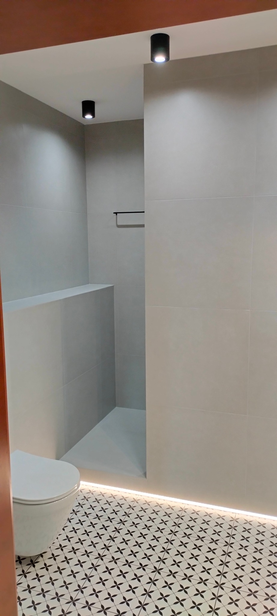 Nowoczesna łazienka z szarymi płytkami ściennymi, wzorzystą podłogą, podświetleniem LED przy podłodze i wiszącym wieszakiem na ręczniki.