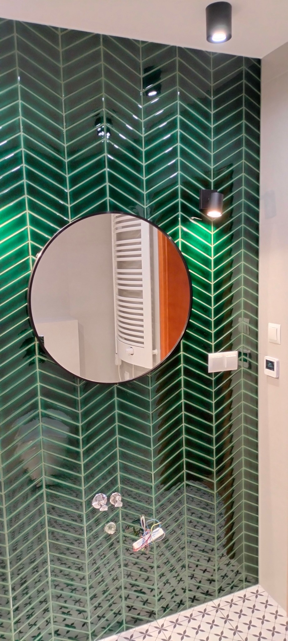 Ściana łazienki wyłożona błyszczącymi, zielonymi płytkami w jodełkę, z okrągłym lustrem, baterią i grzejnikiem widocznym w odbiciu. Na ścianie czarna lampa punktowa i białe włączniki.