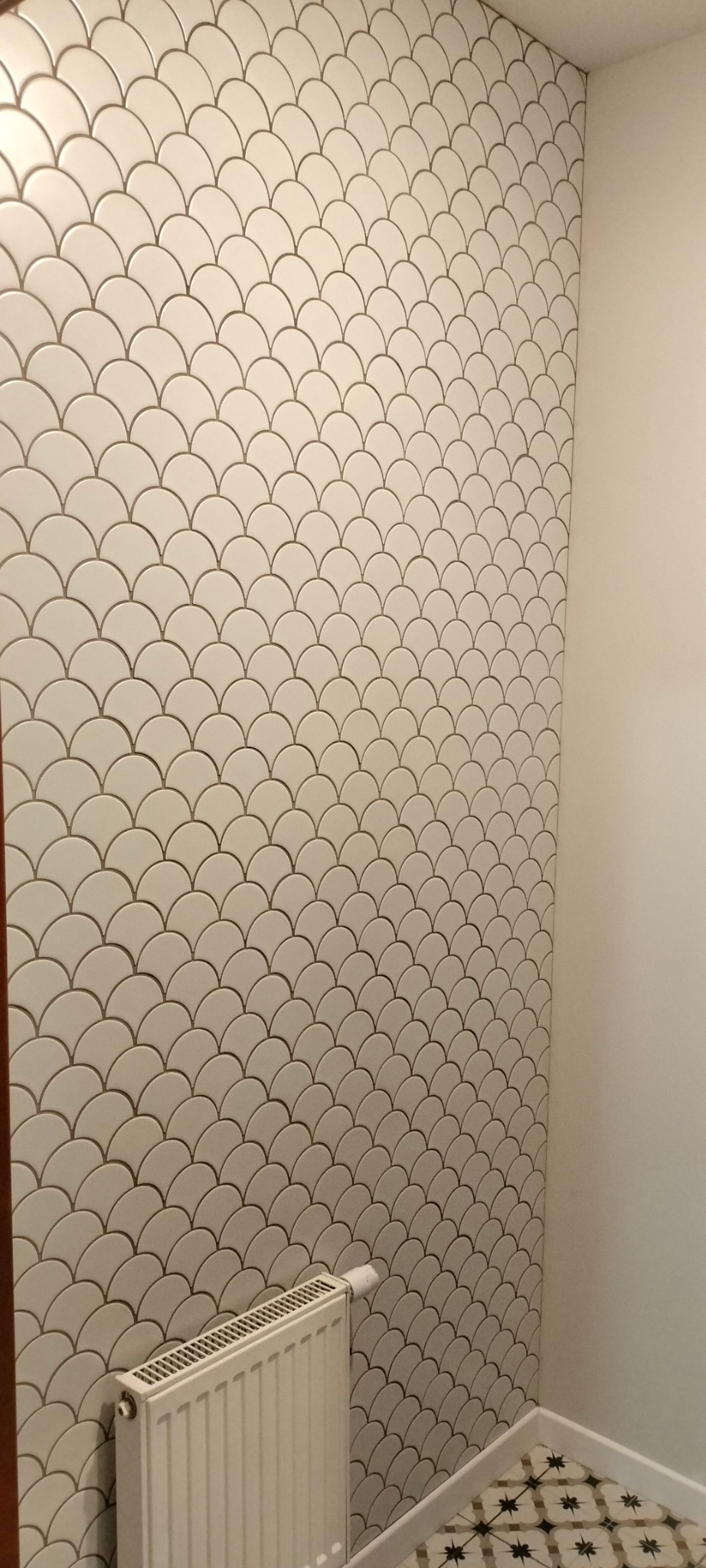 Ściana wyłożona białymi płytkami w kształcie rybiej łuski z szarą fugą, widoczny grzejnik i fragment podłogi z wzorem geometrycznym w łazience.