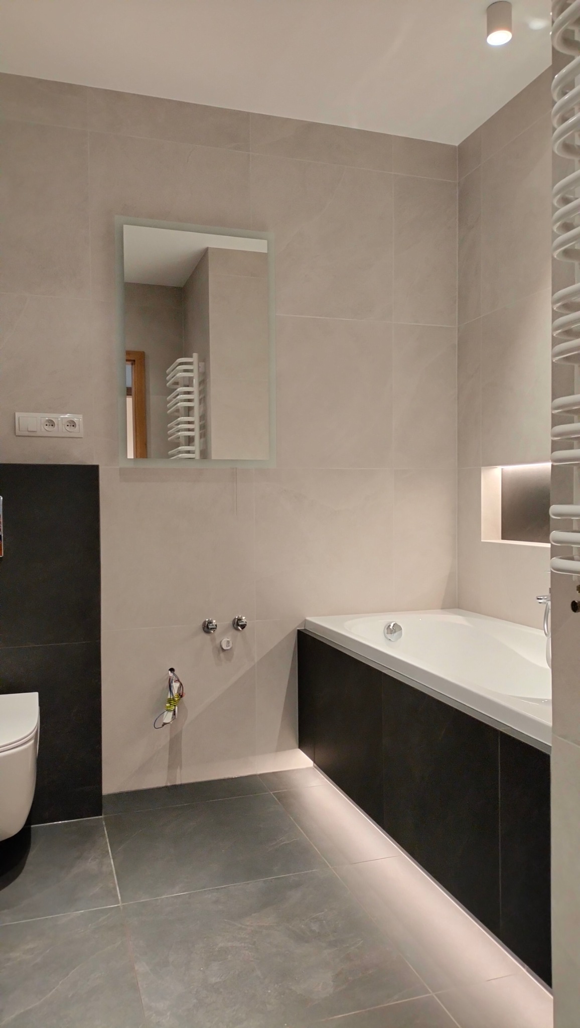 Nowoczesna łazienka w odcieniach szarości, z podświetlaną wanną, dużymi płytkami na ścianach i podłodze, oraz prostokątnym lustrem z podświetleniem.