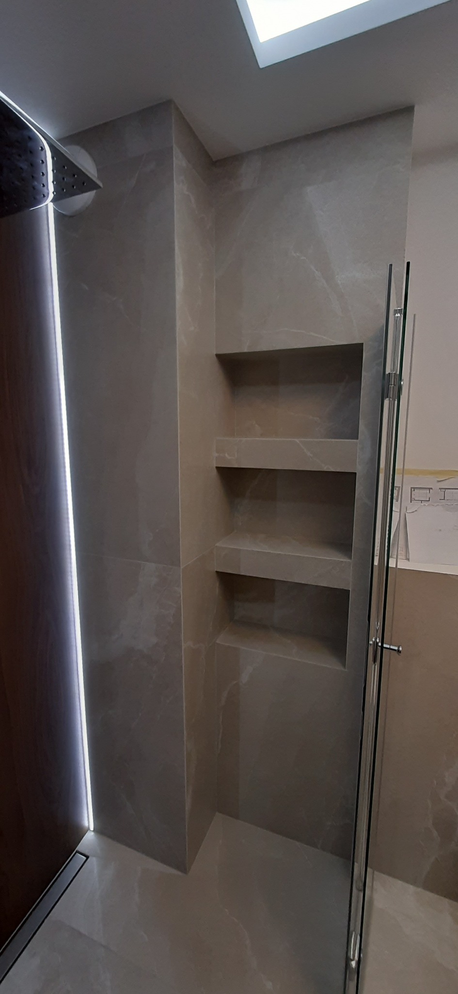 Wykończona łazienka z szarymi płytkami imitującymi marmur, wbudowane półki w ścianie, odpływ liniowy i szklana kabina prysznicowa.