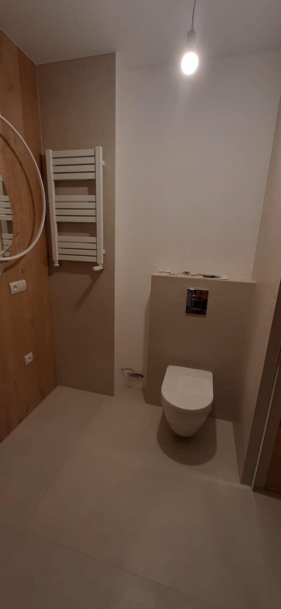 Nieduża łazienka w jasnych beżach z wiszącą toaletą, grzejnikiem drabinkowym i okrągłym lustrem z oświetleniem górnym