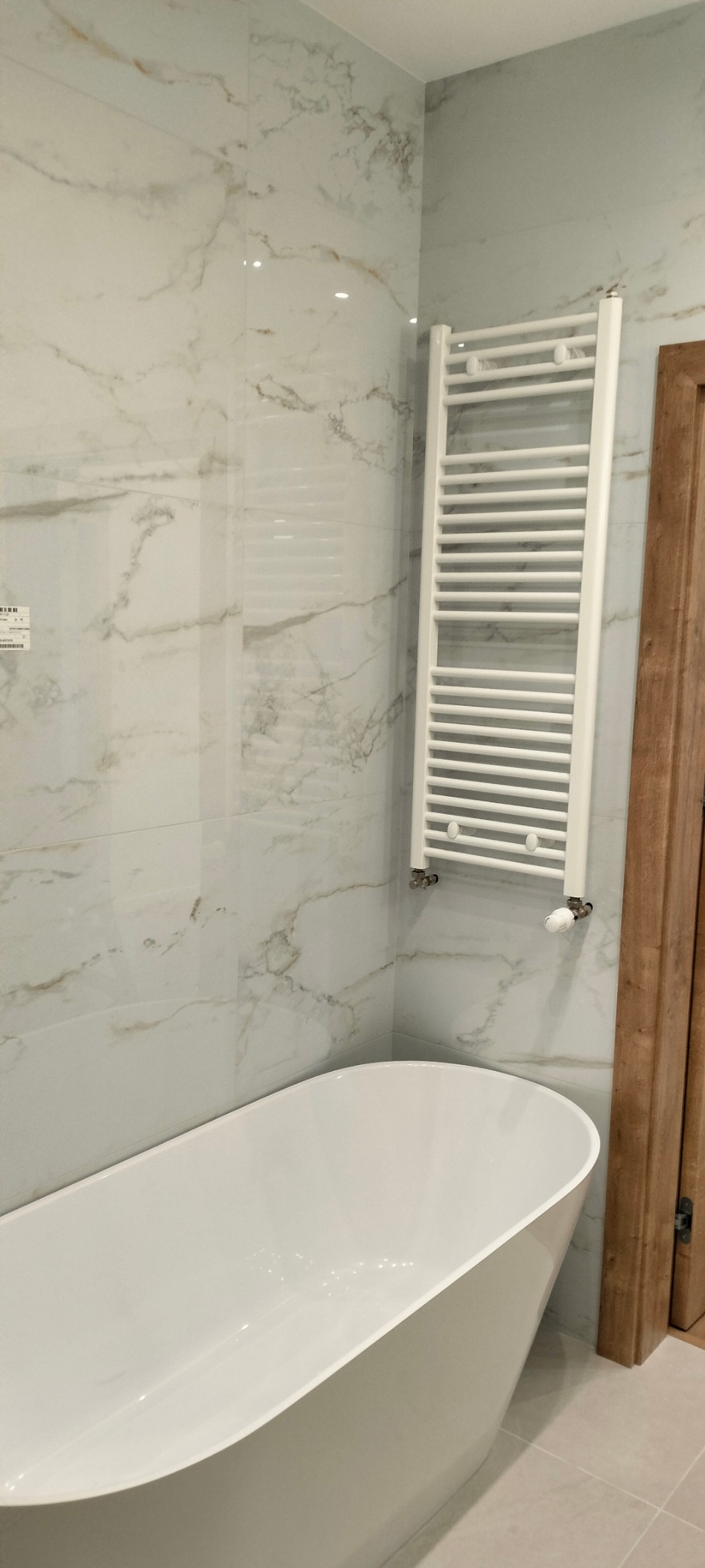 Biała wanna wolnostojąca w łazience z marmurowymi płytkami na ścianach i grzejnikiem drabinkowym.