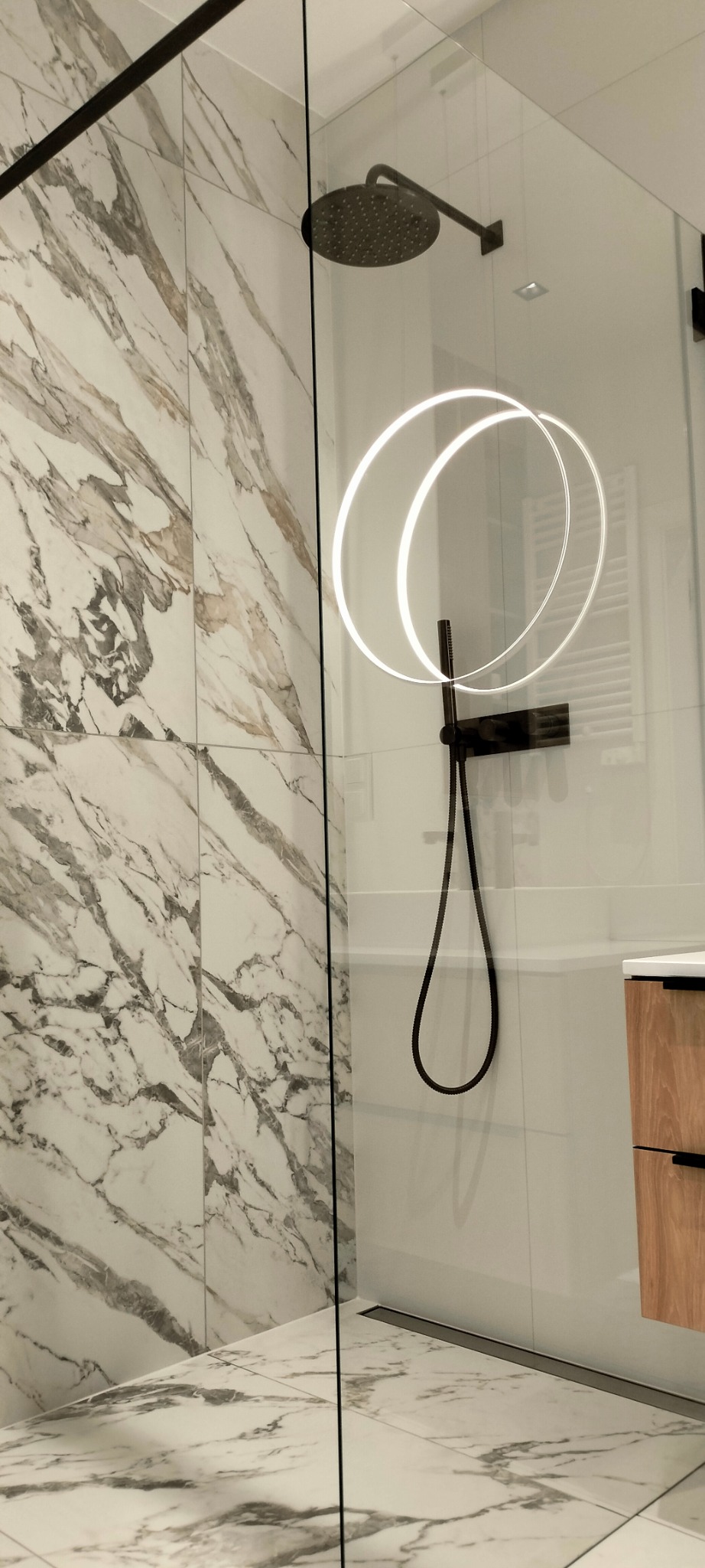 Nowoczesna kabina prysznicowa z czarną armaturą, deszczownicą i marmurowymi płytkami na ścianach i podłodze, oświetlenie LED w kształcie okręgów na szklanej ściance.