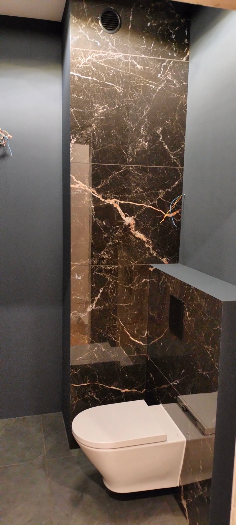 Nowoczesna toaleta podwieszana w łazience wykończonej czarnymi płytkami imitującymi marmur z jasnymi żyłami, widoczne wystające przewody elektryczne.