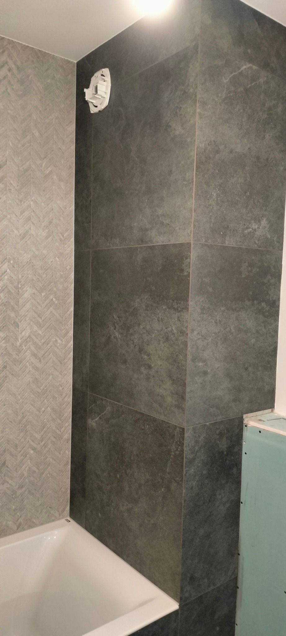 Częściowo wykończona łazienka z szarą, chevronową ścianą i ciemnymi, marmurkowymi płytkami na ścianie prysznicowej, widoczna biała wanna i fragment ściany z płyt kartonowo-gipsowych.