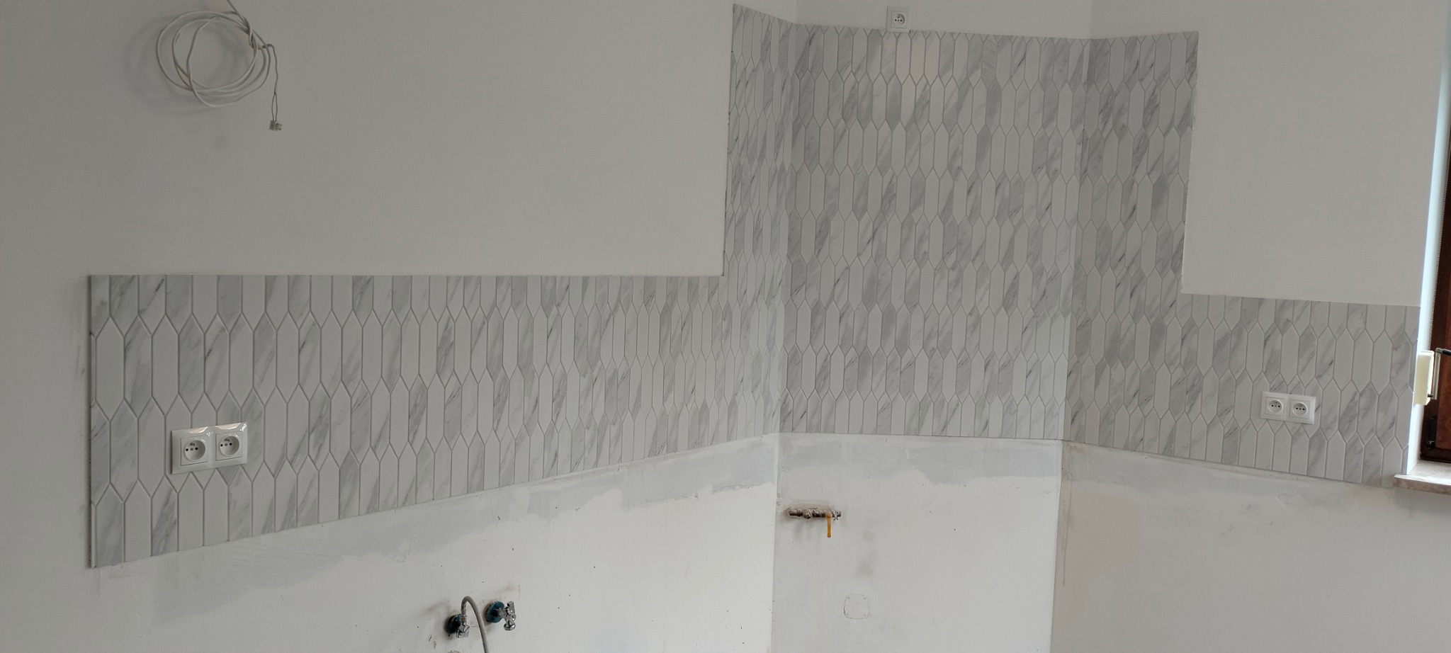 Ściana w trakcie układania płytek o wzorze marmuru, widoczne gniazdka elektryczne i wystające kable, ślady zaprawy na ścianie.