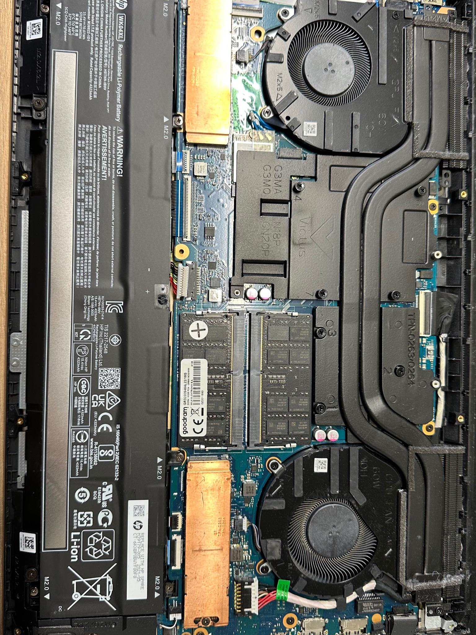 Wnętrze laptopa HP Victus z widoczną baterią, dwoma wentylatorami chłodzącymi, modułami RAM Goodram, płytą główną oraz układem chłodzenia z rurkami cieplnymi.
