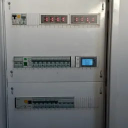 Rozdzielnia elektryczna w metalowej obudowie z bezpiecznikami, wyłącznikami i modułami sygnalizacyjnymi z diodami LED. Widoczne oznaczenia i numeracja.