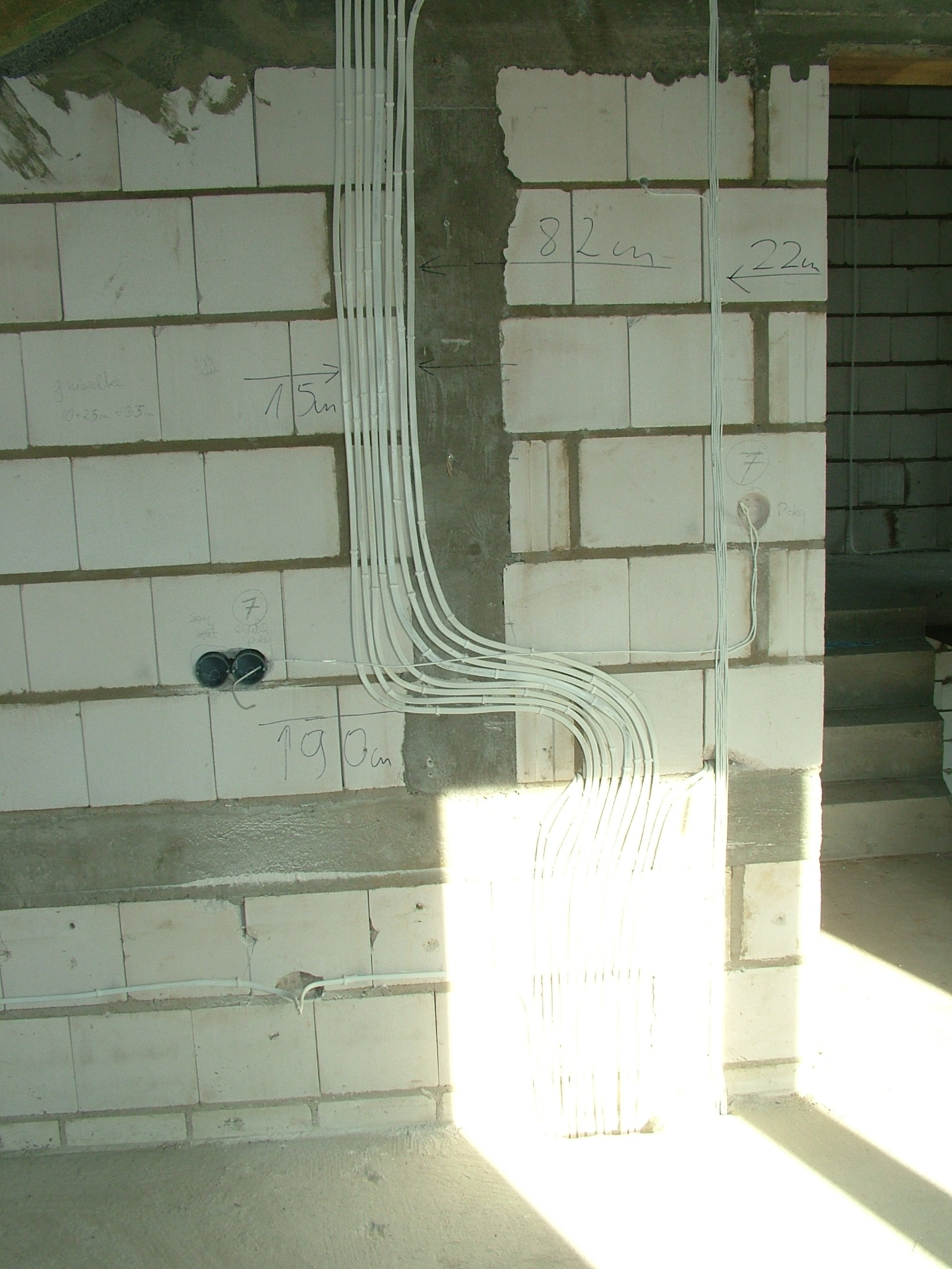 Wewnętrzna instalacja elektryczna w ścianie z białych bloczków, z widocznymi przewodami w peszlach i zaznaczonymi wymiarami na ścianie. Etap prac wykończeniowych.